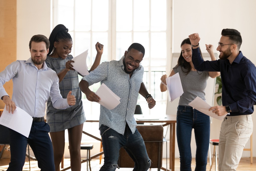 Excited multiracial millennial colleagues dance holding paperwork happy with business project success or good work results, overjoyed diverse employees have fun in office celebrating goal achievement