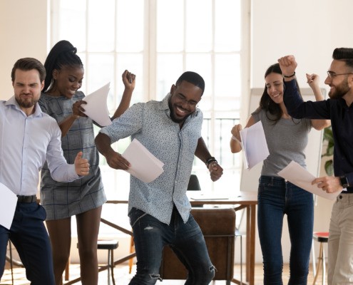 Excited multiracial millennial colleagues dance holding paperwork happy with business project success or good work results, overjoyed diverse employees have fun in office celebrating goal achievement