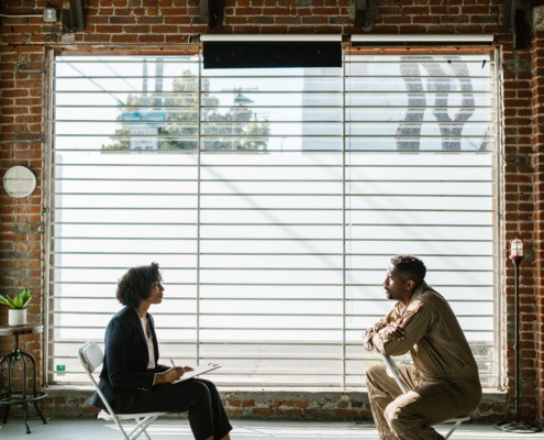 Decorative image of woman and man talking inside in front of a large window, both on chairs. pic by Rodnae Productions courtesy of pexels.